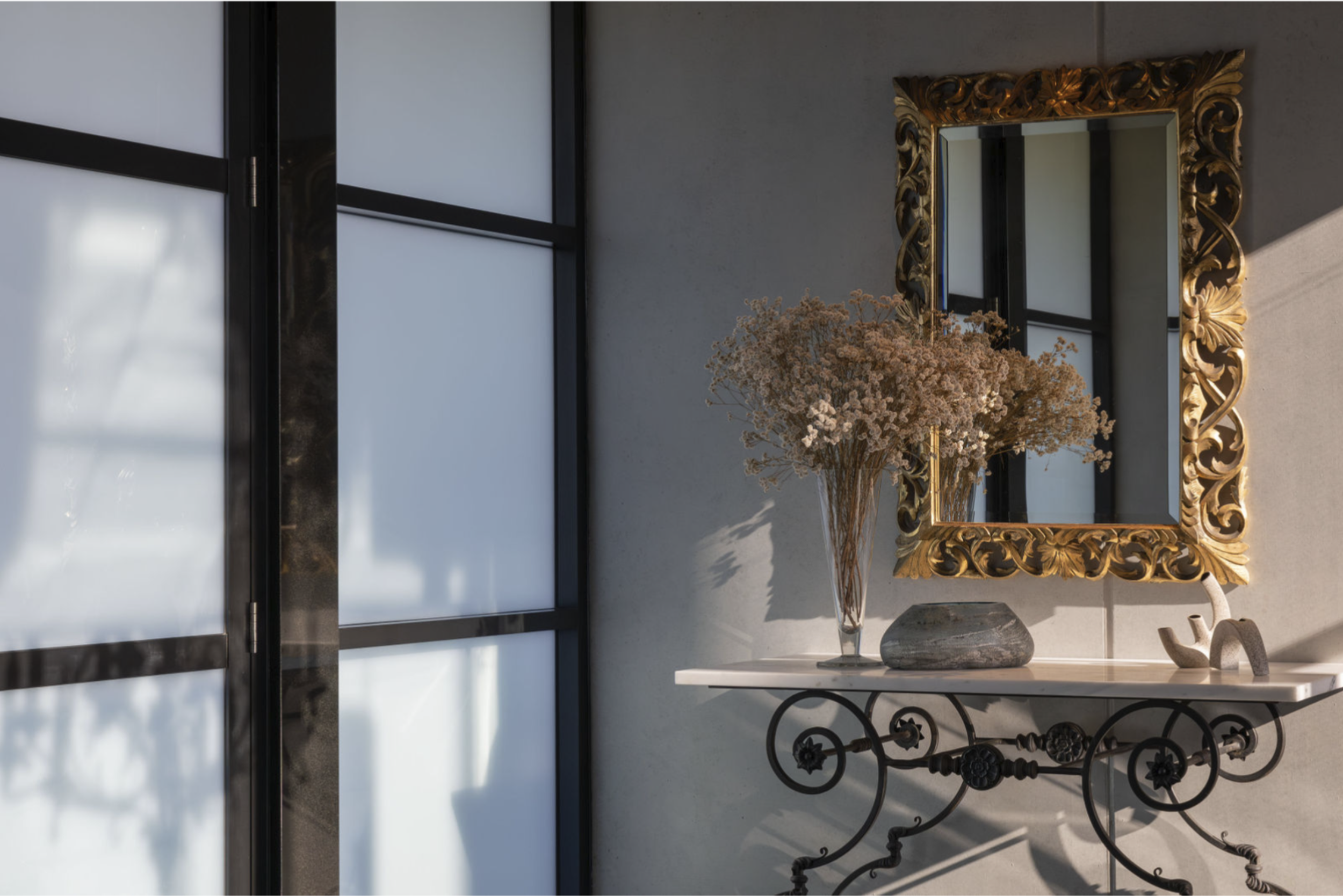 A decorative interior wall with a large ornate gold-framed mirror, a white marble-top console table with wrought iron legs and a glass vase of dried pink flowers