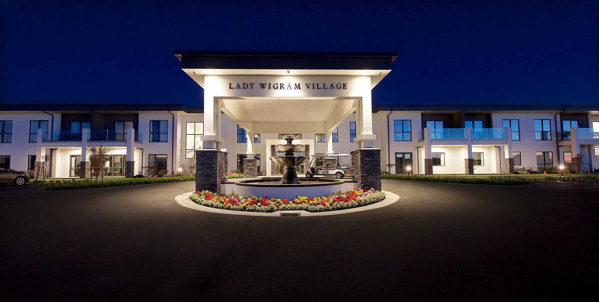 Night view of Lady Wigram Village apartment entrance with a lit fountain, surrounded by flowers and modern buildings