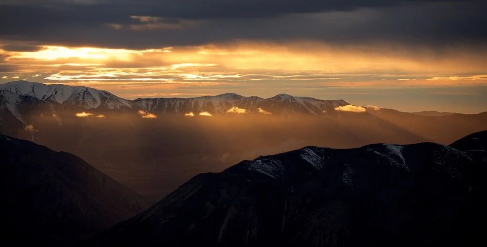 Sunset over a mountain range with dark clouds, illuminated clouds and snow-capped peaks in the background